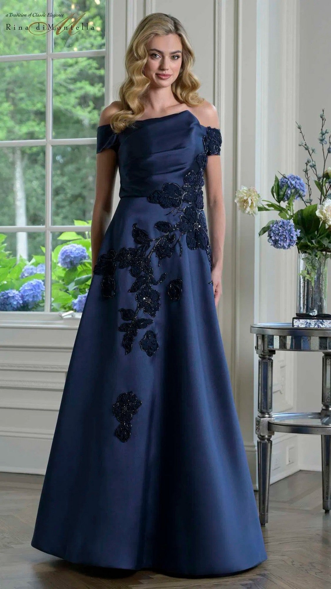 A woman stands indoors wearing the Rina Di Montella RD3344 long formal dress in navy; blue hydrangeas sit on a table beside her.